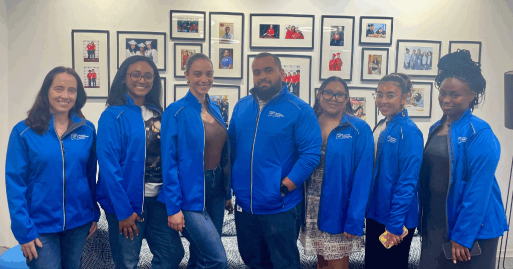 Seven people, including Benjamin, wear matching blue jackets stand side by side indoors, posing for a group photo in front of a wall with framed pictures.