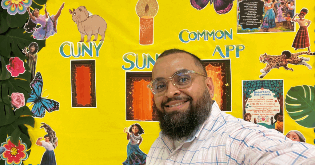 Benjamin smiles in front of a colorful bulletin board decorated with animated characters and college application information.