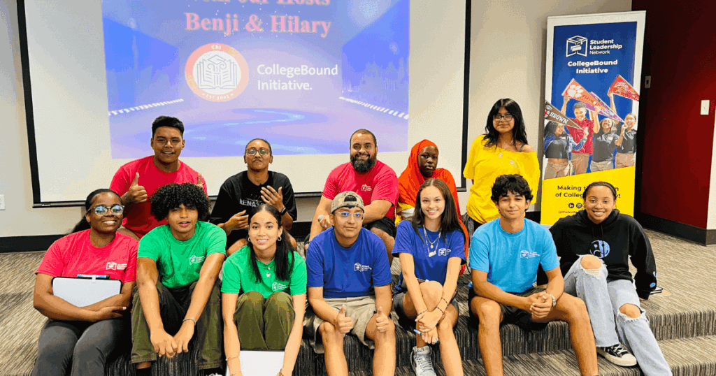 Benjamin sits with students and adults wearing colorful t-shirts, all posing together in front of a CollegeBound Initiative presentation screen in a classroom setting.